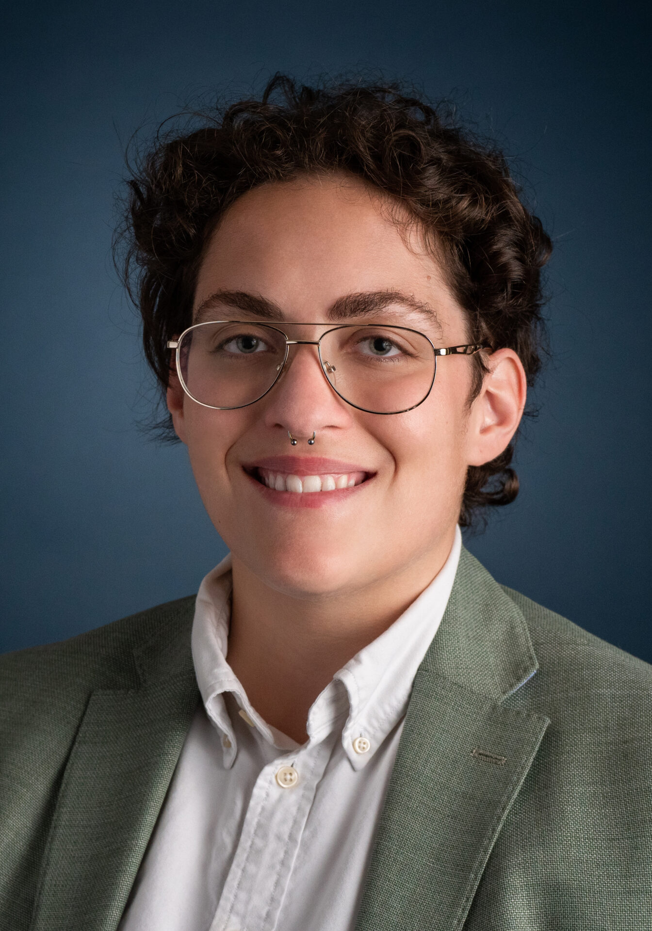 Stevie Mays is a white person with short blow curly hair, they are smiling wearing glasses and are in a professional white button up collared shirt with a mint green suit. They are behind a navy blue background